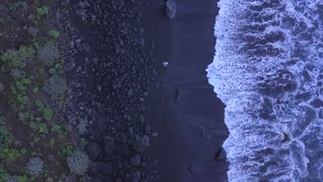 man walking along waves swashing on volcanic dark sand beach, aerial