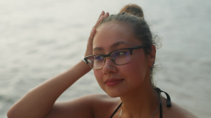 Donna felice che si gode il tramonto in riva al mare. Giovane donna con gli occhiali che sorride sulla riva durante il tramonto. Donna allegra con collana e occhiali che irradia felicità vicino all'oceano durante l'ora d'oro