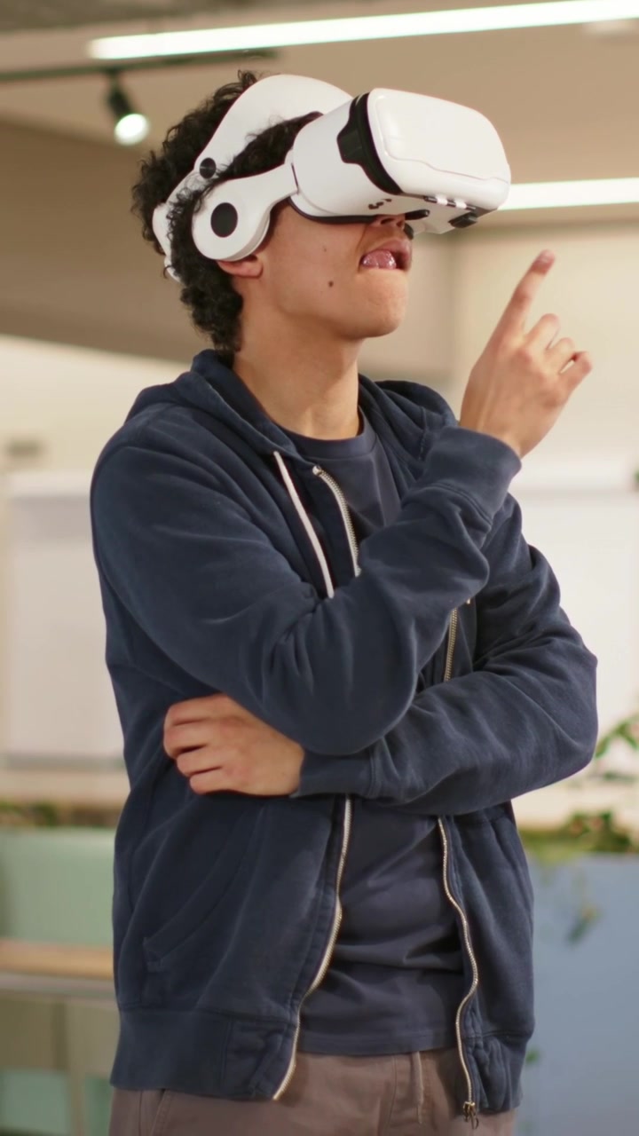 Video verticale: Uomo che attiva le cuffie, con visore VR bianco e felpa con cappuccio blu scuro, che testa l'interfaccia VR in ufficio.
