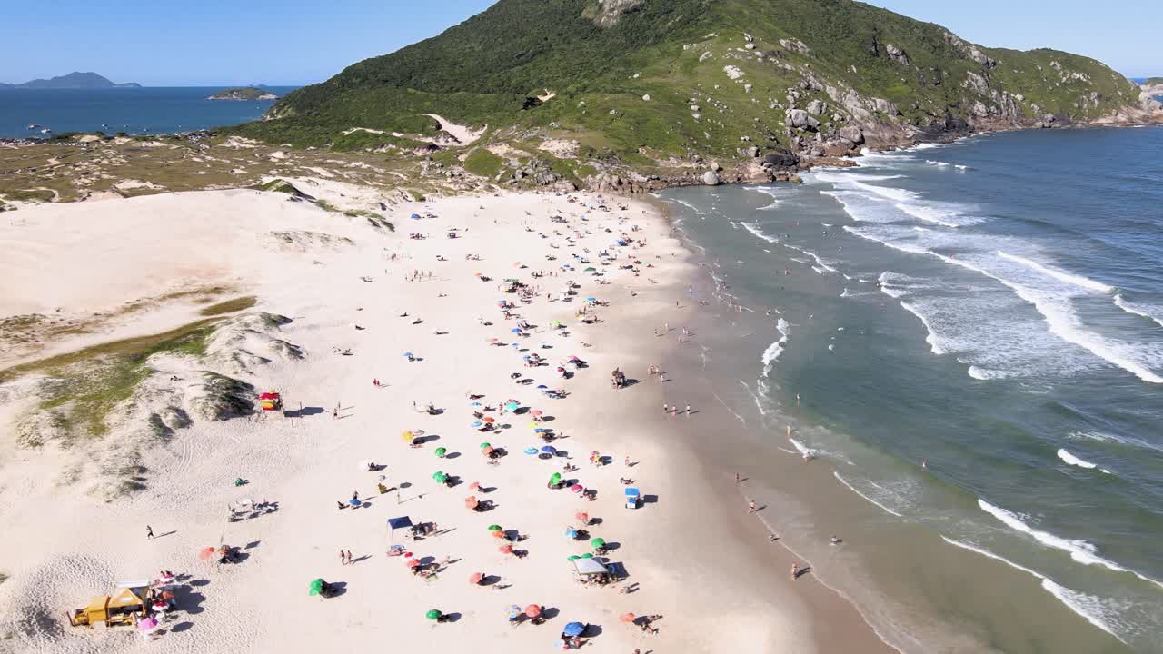Elokuvamainen laaja ilmakuva Santinhon rannasta Florianópolisissa, Brasiliassa, jossa näkyy tungosta ranta, valkoisia hiekkadyynejä ja Morro dos Ingleses -vuori kirkkaan sinisen taivaan alla.