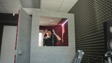 a man dressed in black stands in front of a studio microphone, wearing headphones as he sings in a red soundproofed room