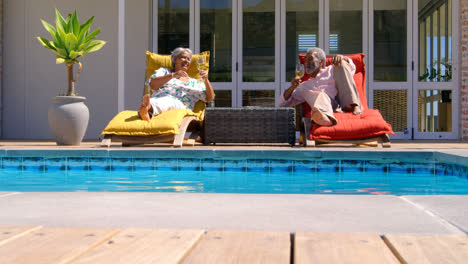 Vista-Frontal-De-Una-Pareja-De-Ancianos-Negros-Relajándose-En-Una-Tumbona-En-La-Piscina-En-El-Patio-Trasero-De-Su-Casa-4k