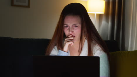 anxious woman sitting on sofa at home at night looking at laptop concerned about social media or bad news 3