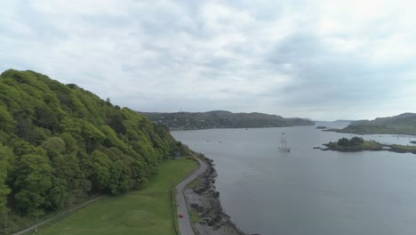 Toma-Aérea,-Acercándose-A-La-Bahía-De-Oban-Desde-Dunollie-Woods,-Siguiendo-La-Carretera-Que-Bordea-El-Mar.