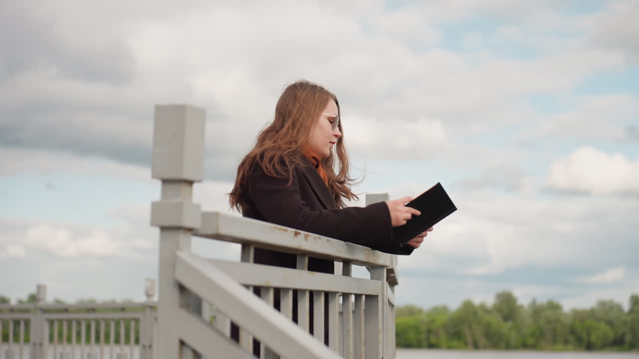 Student with long hair wearing winter jacket walks outdoors reading book near metal railing under cloudy sky, showing focus, intelligence, and curiosity while enjoying peaceful learning atmosphere