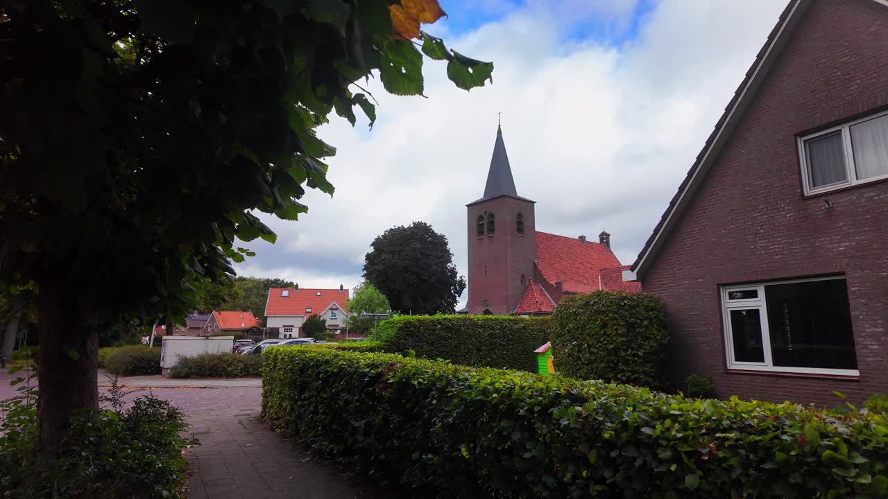 Brick church with pointed tower rises above hedges, trees, and surrounding houses in a quiet Ommen neighborhood. Ommen, Overijssel, Netherlands (Ommen, Overijssel, Nederland)