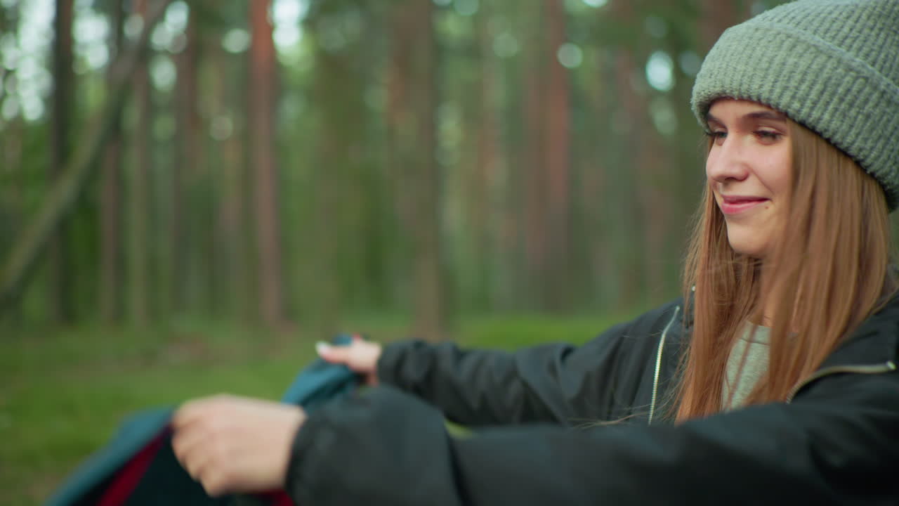 Close up of lady with long hair wearing green beanie and black jacket smiling while holding green tent fabric, blurred forest background filled with trees