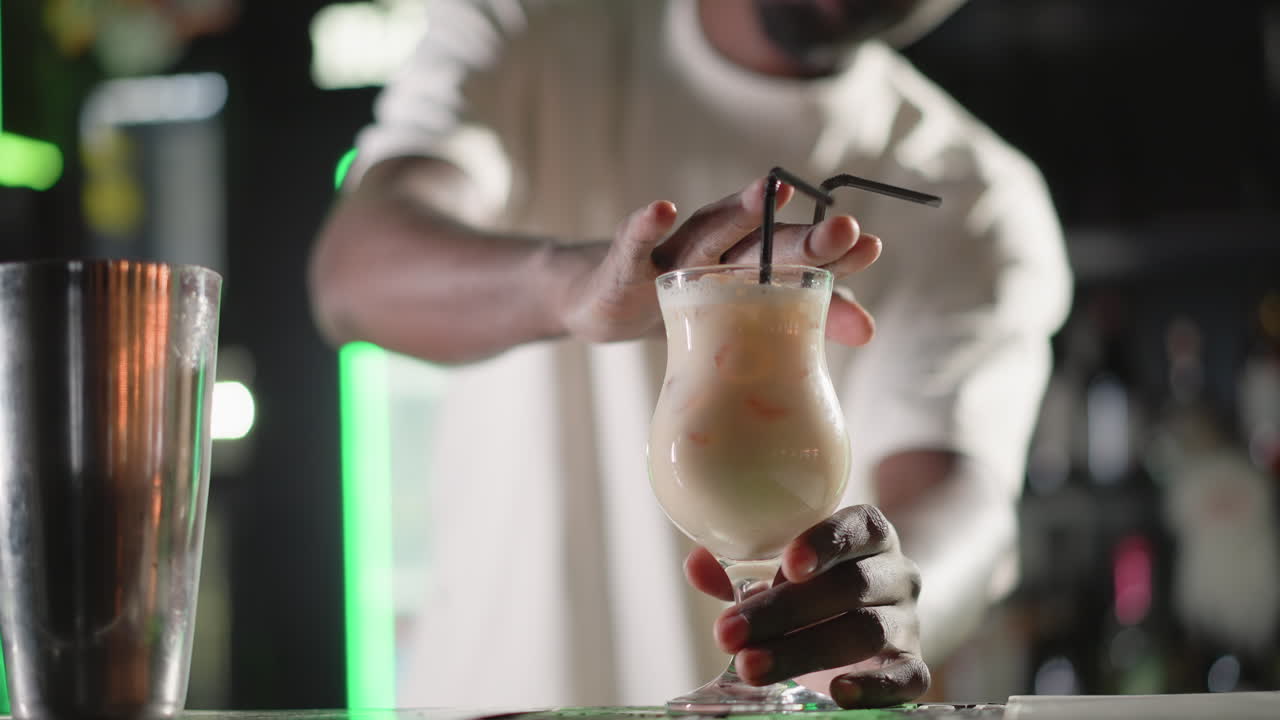 Bartender adding straw to cocktail in elegant glass, preparing to serve drink. Behind, illuminated Heineken neon sign and well-stocked bar shelves enhance vibrant bar atmosphere