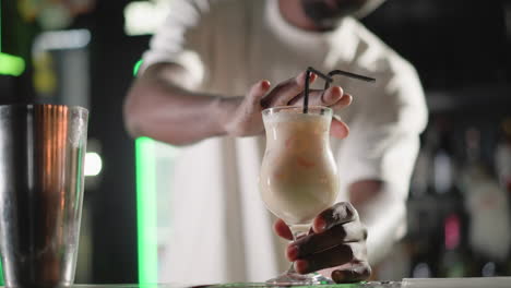 bartender adding straw to cocktail in elegant glass, preparing to serve drink. behind, illuminated heineken neon sign and well-stocked bar shelves enhance vibrant bar atmosphere