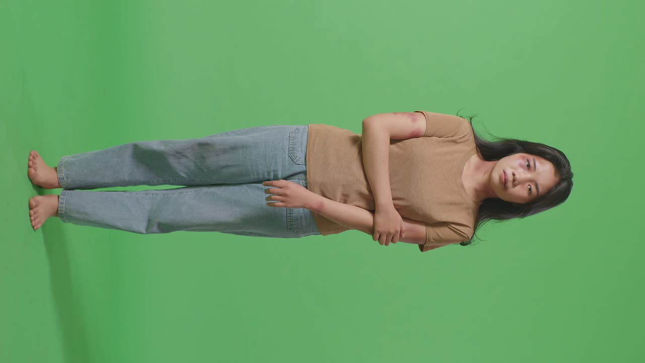 얼굴에 이 있는 폭력의 희생자인 젊은 아시아 여성의 몸은 그녀의 팔을 들고 녹색 스크린의 배경 스튜디오에서 슬프게 카메라를 쳐다보고 있습니다.