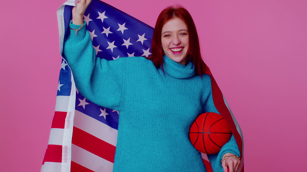 una joven pelirroja fanática de baloncesto sosteniendo la bandera de los estados unidos haciendo el gesto del ganador, bailando sola