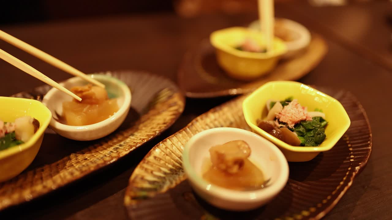 温かい照明の下、小さな陶器の器から箸で食べ物をつまんでいるクローズアップショット。