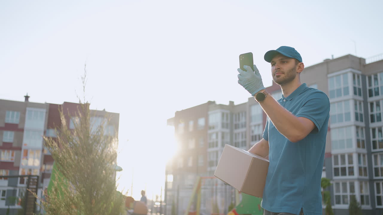 The postman with glasses carries the parcel and looks at the delivery address via mobile phone. search for the address of the delivery customer. Delivery guy with a cap and a box