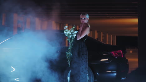 mujer elegante con flores por un coche en un garaje de estacionamiento por la noche