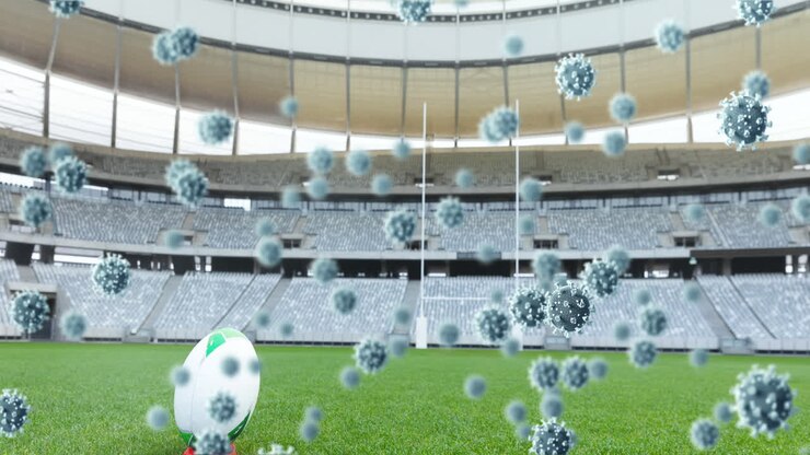 Molteplici cellule Covid-19 che fluttuano contro una palla da rugby su uno stadio sportivo