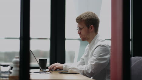 Junger-Mann-Mit-Brille-Und-Lächeln,-Während-Er-An-Seinem-Laptop-Arbeitet,-Um-Frühmorgens-Bei-Seiner-Tasse-Kaffee-Alle-Seine-Geschäfte-Zu-Erledigen