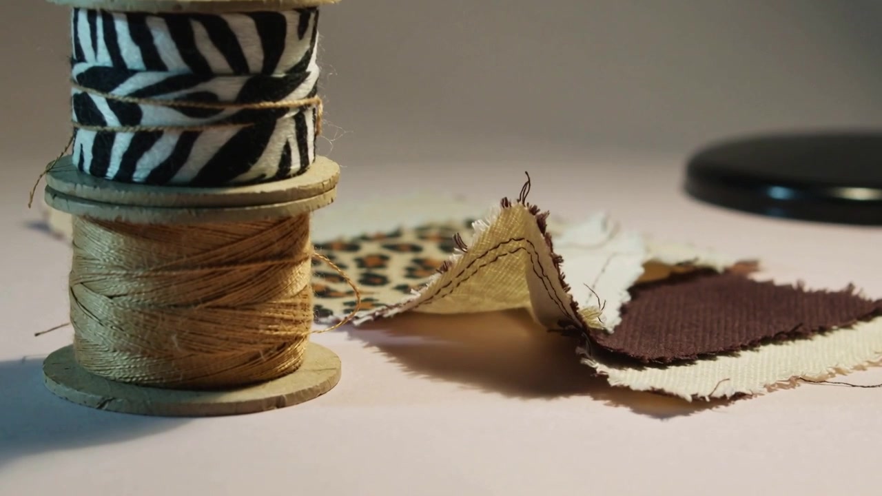 De camera beweegt en toont gestapelde spoelen met zebra lint, een genaaid staaltje bij een lamp op een knutseltafel, met zichtbare naden.