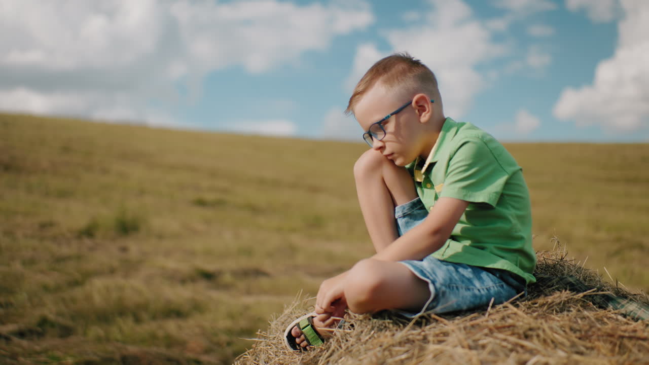 el niño se sienta en una bola de heno con la cabeza inclinada, perdido en sus pensamientos, rodeado de un vasto campo abierto bajo un cielo azul brillante, capturando un momento tranquilo y pacífico en el campo caluroso de un día soleado.