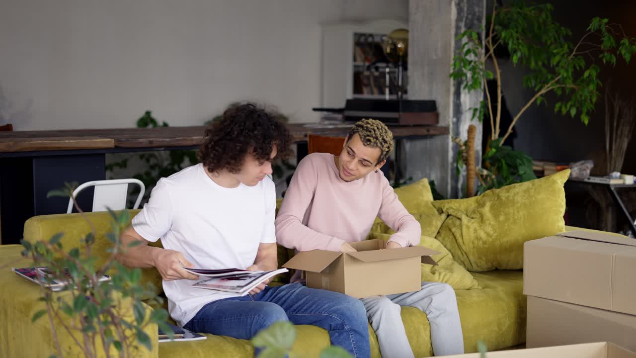 Smiling gay couple move to a new apartment, unpacking boxes while sitting on sofa