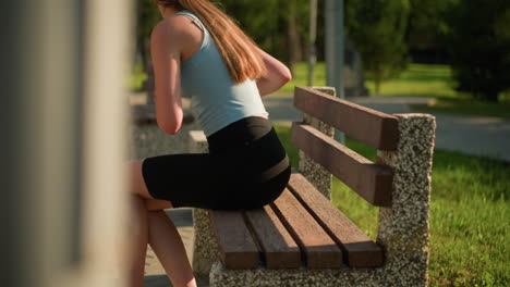 side view of young lady placing cyan roller skates on wooden bench under sunlight, and has a black wristband on right hand, background features lush greenery, trees, and nearby benches