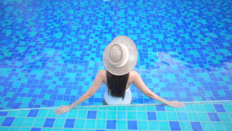 De-Espaldas-A-La-Cámara,-Una-Mujer-Con-Un-Traje-De-Baño-Blanco-De-Una-Pieza-Y-Un-Sombrero-De-Paja-Se-Recuesta-Sobre-Sus-Brazos-Mientras-Observa-La-Piscina.