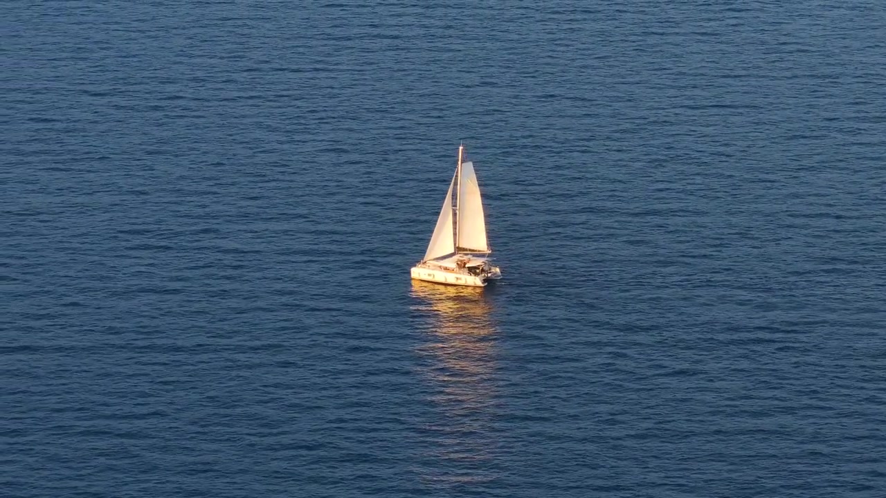 Um veleiro é visto navegando na água. A vela está içada, pegando vento. O barco está em uma área ampla e aberta do mar. A luz do sol reflete na superfície da água.