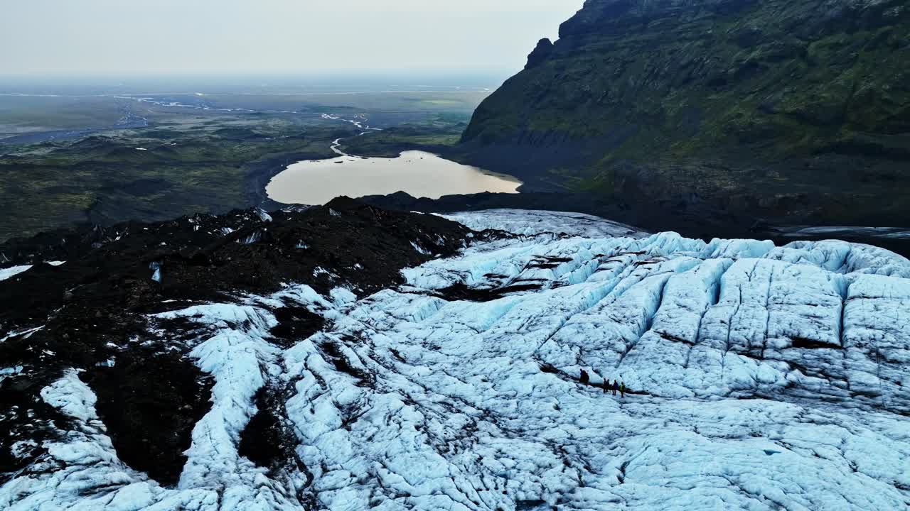 En drone svæver hen over en gletsjertunge med sprækker, der er stribet med mørk vulkansk aske, og åbner sig mod en fjern floddal og sø under stejle bjergskråninger.