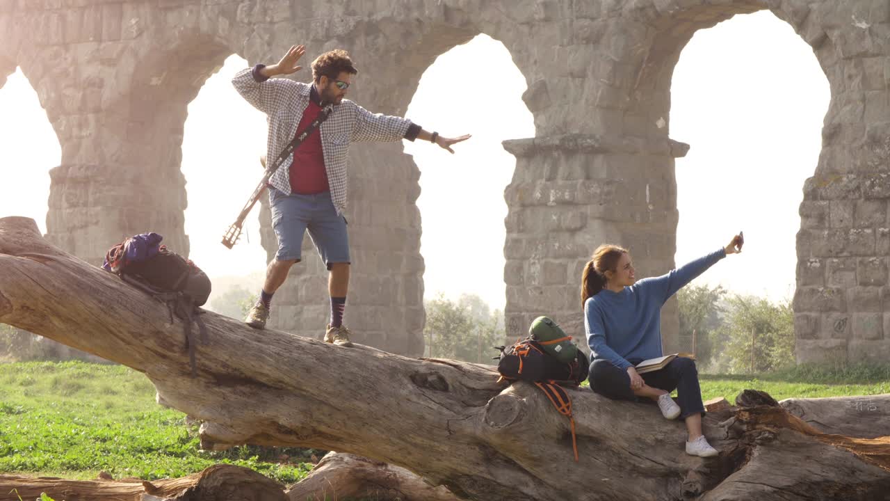 una pareja feliz de mochileros turistas en un tronco de troncos tomando selfies fotos con el teléfono inteligente frente a las antiguas ruinas del acueducto romano en el romántico parque parco degli aquedotti en roma al amanecer brumoso trípode de cámara lenta