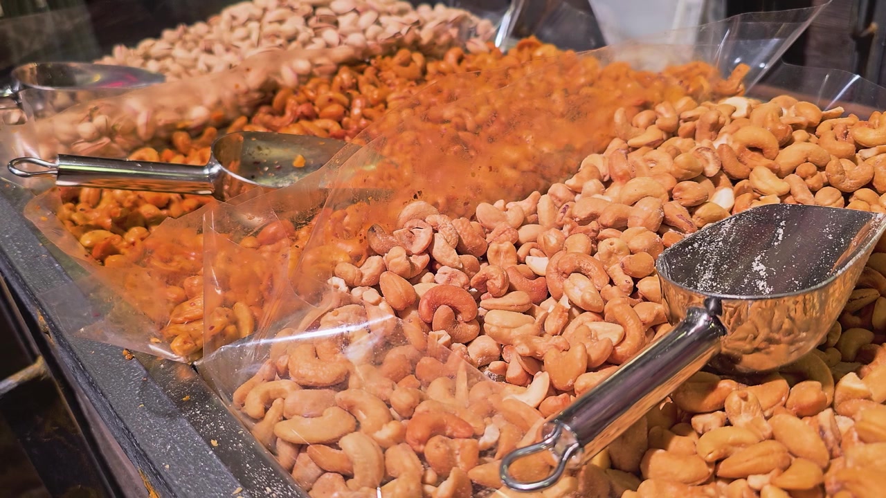 Nærbilde over rader med ristede nøtter og snackblandinger i en markedsdisk, med reflekterende metallskjeer, gjennomsiktige beholdere, varme lys og teksturert presentasjon av bulkmat i butikk