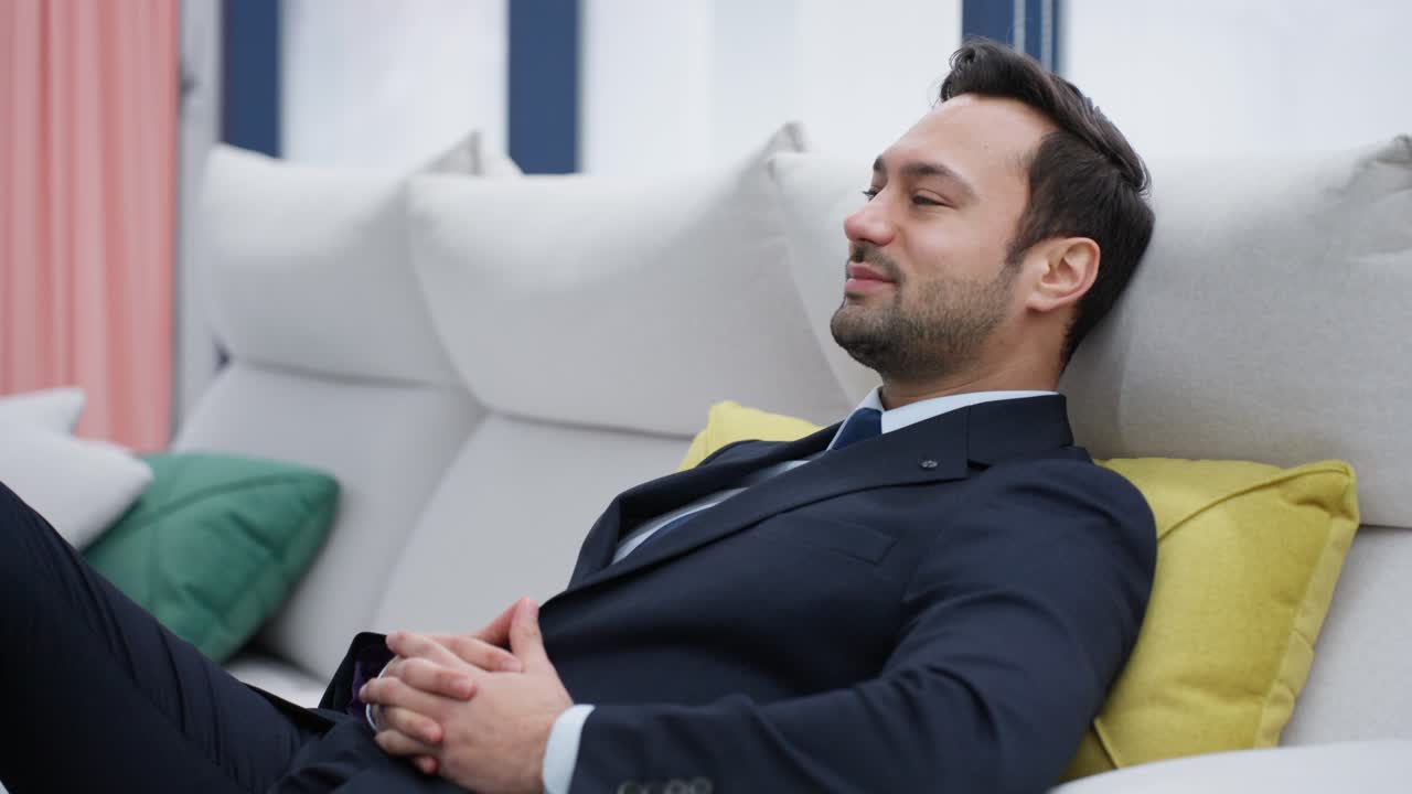 An inspired dreaming man in a formal suit is reclining on a modern white couch with his hands clasped, businessman pensive guy employer relaxing in a positive mood with a satisfactory smile confidence