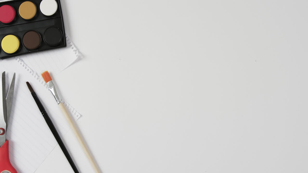 Overhead view of paints, brushes and scissors on white background with copy space, in slow motion
