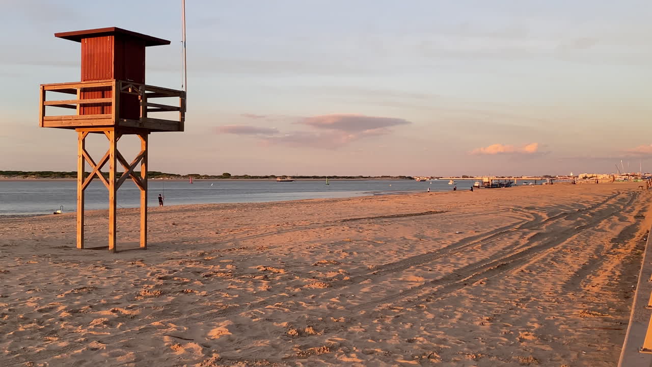 Una torre de vigilancia de madera se alza sobre la playa vacía de Sanlúcar de Barrameda bajo la cálida luz del atardecer junto a la tranquila desembocadura del río