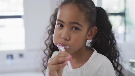 Portrait-of-happy-biracial-girl-looking-at-camera-and-brushing-teeth