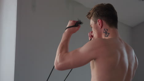 shirtless man with neck tattoo performing resistance rope exercise near large window in minimalist indoor setting, showcasing upper body strength and muscular back during intense training routine