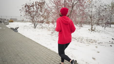 back view of woman jogging along paved snow-covered path with a warm smile during winter, surrounded by serene park scenery and barren trees, a pigeon flies nearby as she approach