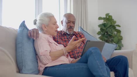 happy senior biracial couple sitting on couch and using tablet at home, slow motion