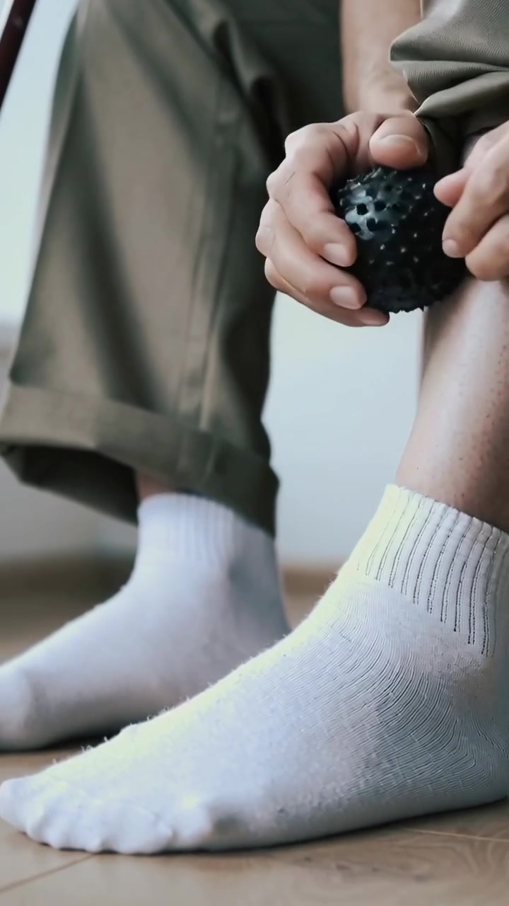 Vertical video: Placing spiky ball on ankle, man pressing rolling for relief at home, cuffed khakis