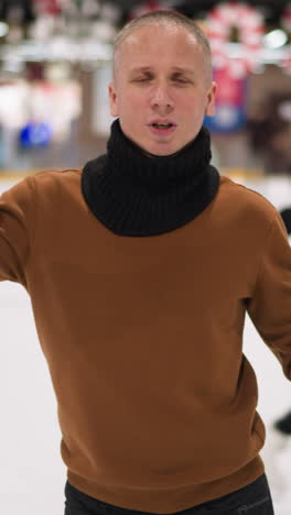 hombre calvo de mediana edad con un suéter marrón y una bufanda negra bailando alegremente en una pista de hielo, apuntando hacia arriba, con patinadores borrosos en el fondo