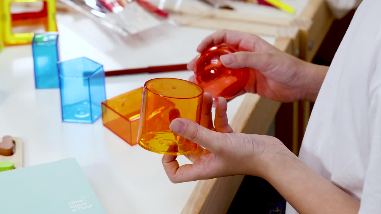 Child arranges translucent geometric solids on desk under bright lighting, demonstrating hands-on mathematics education