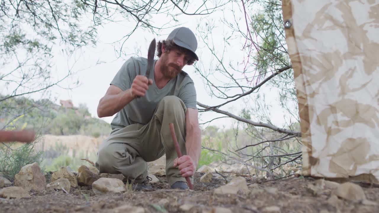 Bearded caucasian male survivalist banging stick into ground with machete at camp in wilderness