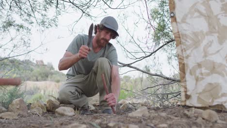 bearded caucasian male survivalist banging stick into ground with machete at camp in wilderness