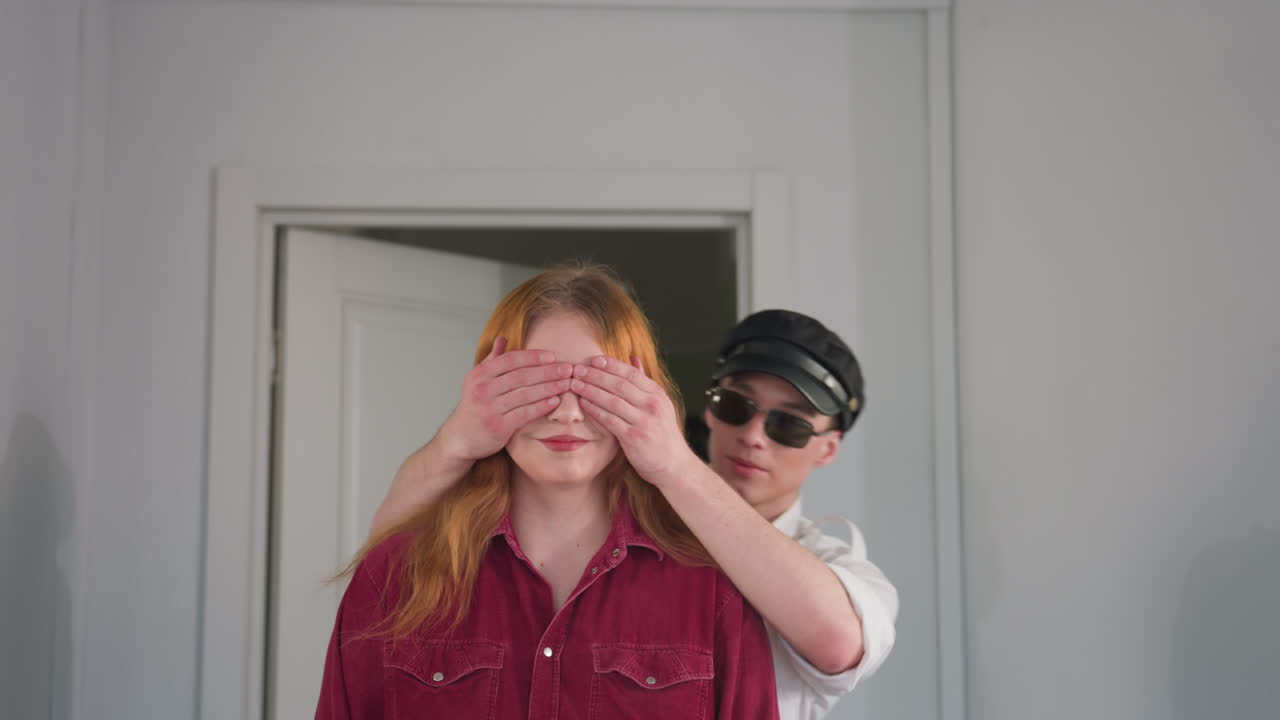Playful moment as man in cap and sunglasses gently covers eyes of young woman in red shirt from behind, standing together in doorway with soft lighting and calm expressions