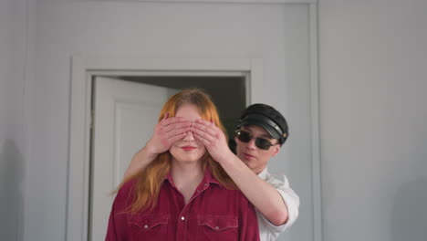 playful moment as man in cap and sunglasses gently covers eyes of young woman in red shirt from behind, standing together in doorway with soft lighting and calm expressions