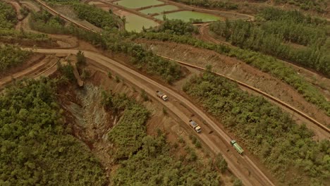 Trucks-driving-on-a-dusty-track-through-Taganito-nickel-Mines,-Philippines,-surrounded-by-trees-and-industrial-land