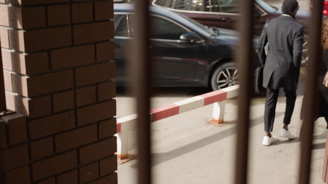 Two students wearing long winter jackets meet at hallway entrance, walking opposite directions past block rails, sunlight on hair, parked cars beyond and candid campus vibe with gentle breeze
