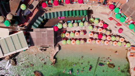 An-overhead-aerial-of-colorful-umbrellas-and-tourists-along-the-beaches-of-Marina-Piccola-on-Capri,-a-famous-island-that-is-a-popular-luxury-vacation-destination-in-Italy-along-the-Amalfi-Coast