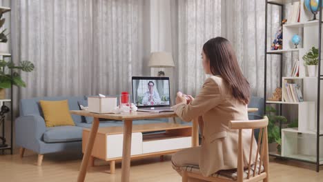 sick asian businesswoman holding a medicine to the video call with doctor on a laptop while working at home