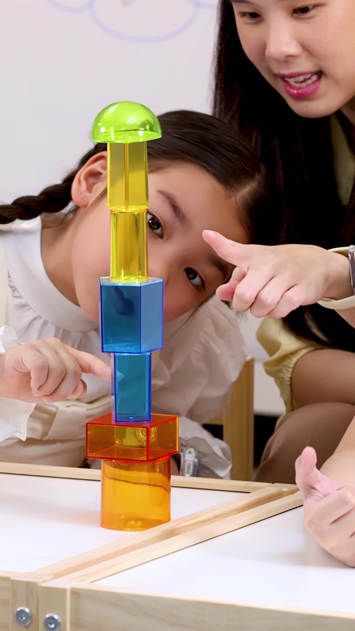 Smiling teacher guides focused child stacking colorful translucent blocks in bright classroom, static close-up shot