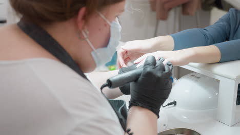 close-up of nail technician focused on filing client nails using electric file, wearing black gloves, face mask, and apron, in well-lit salon setting with client hand resting on padded support