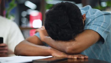 camera capturing teenager in blue shirt lapping elbow with other hand while bending head over desk in soft indoor light conveying thoughtful moment with subtle expression and gentle focus motion zen